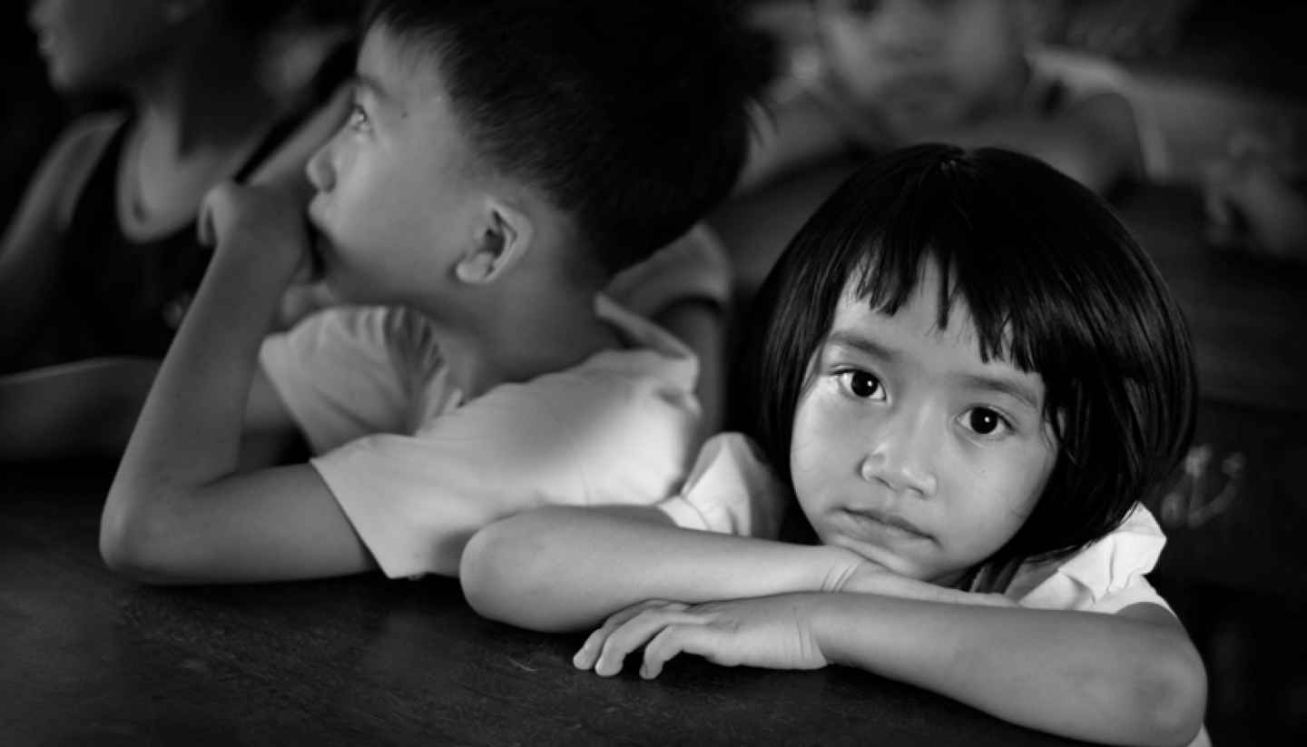 Does voluntourism do more harm than good? - Little girl sitting behind her desk, looking at the camera, surrounded by class mates who are looking in a different direction. Image is in black and white.